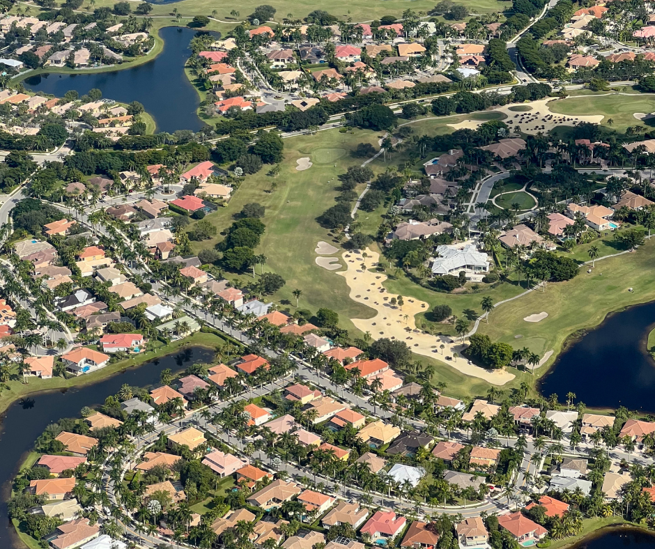 Aerial view of a vibrant suburban community featuring homes with pools, manicured lawns, and curved streets, alongside a dense natural forest at sunset.